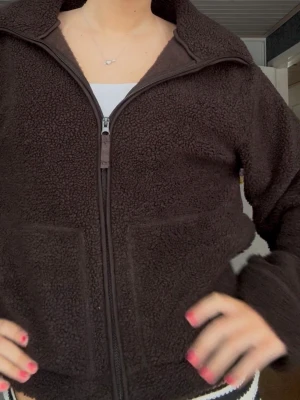 Mörkbrun teddyjacka med dragkedja - Säljer en mysig mörkbrun teddyjacka med hög krage och hel dragkedja framtill. Jackan har två stora fickor på framsidan och är perfekt för kyliga dagar. Materialet är fluffigt och ger en riktigt skön vibe.