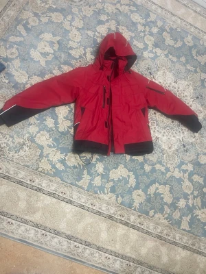 Röd vindjacka från Fristads, M - Säljer en röd vindjacka från Fristads i storlek M. Jackan har huva, svarta detaljer på ärmar och nederkant, samt vita reflexdetaljer. Utrustad med dragkedja, flera fickor och justerbar nederkant. Perfekt för kalla och blåsiga dagar.
