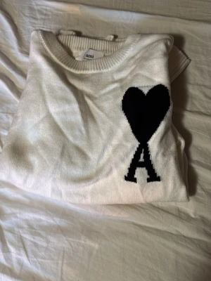 Ami Paris sweatshirt  - Säljer en vit stickad tröja från Ami Paris i storlek M. Tröjan har ett svart hjärta och bokstaven A på bröstet, ribbade muddar och rund hals. Perfekt för dig som gillar stilrena plagg med en twist.
