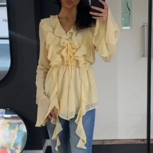 Volangblus - Supersöt ljusgul blus med volanger framtill och längs ärmsluten. Blusen har knytning vid bysten och v-ringning, samt långa, vida ärmar. Tunt och luftigt material som passar perfekt till jeans eller kjol.