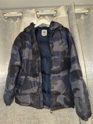 Adidas jacka - Säljer en fet pufferjacka från Adidas med blått och svart camouflage-mönster. Jackan har huva, dragkedja framtill och är vadderad för extra värme. Perfekt för kalla dagar när du vill ha både stil och komfort. Priset är ej hugget i sten