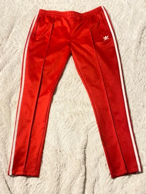 Röda Adidas mjukisbyxor med vita ränder - Säljer ett par röda Adidas mjukisbyxor med vita ränder längs sidorna och diskret Adidas-logga i tyget. Byxorna har resår i midjan, dragkedjeförsedda fickor och klassisk Adidas Trefoil-logga på låret. Materialet är glansigt och känns riktigt sportigt.