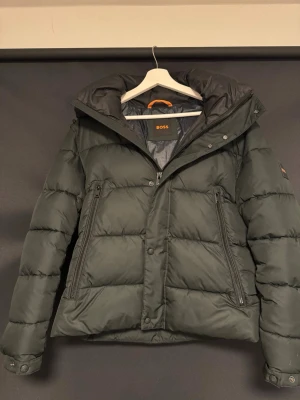 Svart pufferjacka från BOSS - Säljer en svart pufferjacka från BOSS med hög krage och två snedställda fickor med dragkedja. Jackan har en tydlig BOSS-logga på ärmen och stängs med både dragkedja och knappar. Perfekt för kalla dagar och riktigt snygg till streetwear. Använd ett fåtal gånger