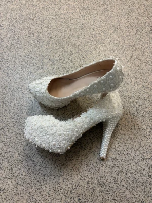 Vita spetsklädda pumps med pärlor - Unika vita pumps täckta med spets och dekorerade med små pärlor. Skorna har hög klack och plattform, samt rund tå. Perfekta för dig som vill sticka ut med eleganta detaljer och romantisk känsla.