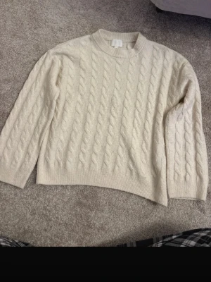 Krämvit kabelstickad tröja - Säljer en krämvit kabelstickad tröja med rund hals och ribbade muddar. Tröjan har ett klassiskt flätmönster och är perfekt för kyligare dagar. Passformen är normal och den är långärmad, vilket gör den både snygg och bekväm.