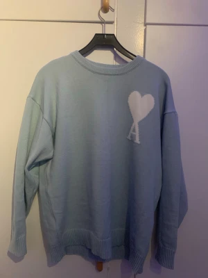 Ljusblå stickad tröja från Ami - Säljer en ljusblå stickad tröja från Ami Alexandre Mattiussi i storlek XL. Tröjan har ett vitt hjärta med bokstaven A på bröstet och ribbade muddar vid ärmslut och nederkant. Perfekt för dig som vill ha en clean och stilren look.