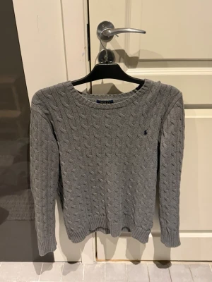 Grå kabelstickad tröja Polo Ralph Lauren - Kabelstickad grå tröja från Polo Ralph Lauren i storlek XL. Klassisk rund halsringning, ribbade muddar och ikoniska broderade loggan på bröstet. Perfekt till lager på lager under höst och vinter. Snygg och tidlös design som alltid funkar.