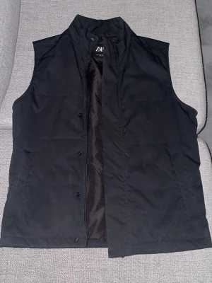 Svart dunväst från Zara - Säljer en svart dunväst från Zara med storlek S, en hög krage och diskreta fickor på sidorna. Västen har dragkedja och tryckknappar framtill, perfekt för lager på lager och enkel att styla till olika outfits.