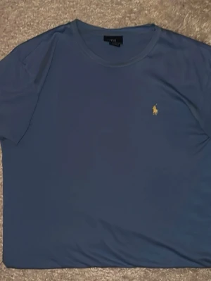 Blå t-shirt från Polo Ralph Lauren - Snygg blå t-shirt från Polo Ralph Lauren med klassisk gul broderad logga på bröstet. T-shirten har rund halsringning och korta ärmar. Tillverkad i mjuk bomull som känns skön mot huden. Perfekt till jeans eller shorts för en clean look.