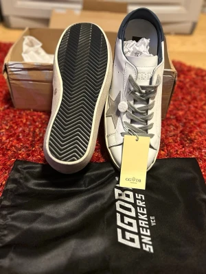 GGDB Superstar vita sneakers stjärna - Golden Goose Superstar sneakers i vitt skinn med grå mockadetaljer och ikonisk stjärna på sidan. Skorna har grå snörning, blå hälflik och beige sula med vintage-look. Kommer med originalpåse och box. Perfekta för dig som gillar streetstyle och premiumkänsla. Priset kan diskuteras 