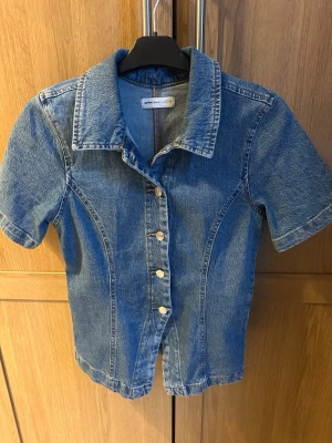 Kortärmad jeanstopp - Säljer en snygg blå jeansjacka från ginatricot i storlek S. Jackan har korta ärmar, klassisk krage och knäppning med fem metallknappar framtill. Knappt aldrig använd .