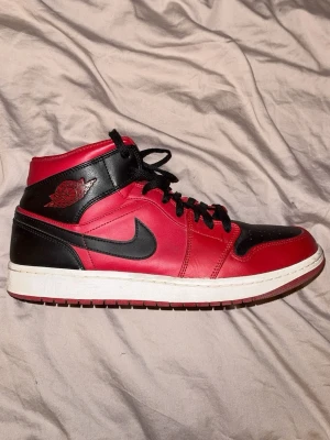 Nike Air Jordan 1 Röd/Svart - Säljer ett par ikoniska Nike Air Jordan 1 i rött och svart skinn med klassisk svart swoosh och snörning. Skorna har vit mellansula och röd yttersula, samt den välkända Air Jordan-loggan på sidan. Perfekta för dig som vill sticka ut med en tidlös sneaker.