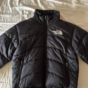 Svart pufferjacka från The North Face - Säljer en svart pufferjacka från The North Face med hög krage och dragkedja framtill. Jackan har två sidofickor med dragkedja och logga på bröstet. Perfekt för kalla dagar och riktigt snygg till streetwear-stilen.