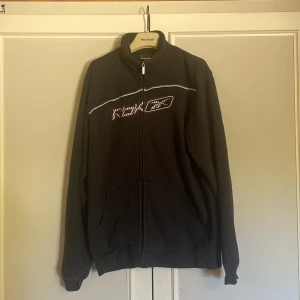 Svart fleecejacka från Reebok - Svart fleecejacka från Reebok med hel dragkedja och hög krage. Jackan har broderad logga på bröstet, två fickor framtill och en vit detalj över bröstet. Perfekt för kyliga dagar och enkel att slänga på sig.
