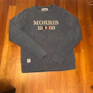 Morris Sweatshirt - Mörkblå stickad tröja från Morris med broderad text 'MORRIS 1968' och flaggdetalj på bröstet. Tröjan har rund hals, ribbade muddar och nederkant samt en liten patch nertill. Perfekt för dig som gillar klassisk och stilren look.