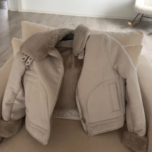 Beige teddyjacka med fuskpäls - Säljer en snygg beige teddyjacka med fuskpälsdetaljer på krage och ärmslut. Jackan har dragkedja framtill och en stilren design som passar perfekt för kyliga dagar.