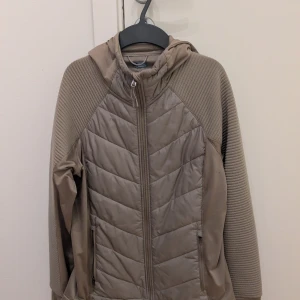 Beige quiltad jacka med huva, M från Hm Move  - Snygg beige jacka med quiltade partier och ribbade ärmar. Jackan har huva, dragkedja framtill och två fickor med dragkedja. Perfekt för kyliga dagar och har en sportig vibe. Materialmix med syntet och polyester för extra komfort.