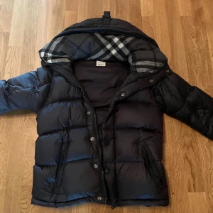 Svart dunjacka från Burberry - Snygg svart dunjacka från Burberry med klassiskt rutigt foder i huvan . Perfekt för kalla dagar och ger en stilren look. Pris kan sänkas vid snabbaffär 