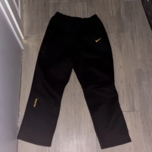 Nocta nike byxor  - Svarta mjukisbyxor från Nike NOCTA med gul Swoosh och NOCTA-logga på benet. Byxorna har snörning i midjan, sidofickor och tryckta detaljer som 'GL' och 'AWR' i gult på benen. Skön passform och stilren look, perfekt för streetwear.