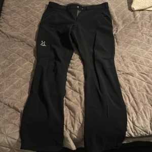 Svarta täckbyxor från Haglöfs herr L - Svarta täckbyxor från Haglöfs i herrmodell, storlek L. Byxorna har en stilren design med dragkedjor vid fickorna och logga på benet. Materialet är slitstarkt och passar perfekt för utomhusaktiviteter under kyliga dagar.