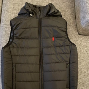 Svart dunväst från Polo Ralph Lauren - Svart dunväst från Polo Ralph Lauren med avtagbar huva och röd logga på bröstet. Västen har quiltad design, två fickor med dragkedja och hel dragkedja framtill. EJ SÄKER OM DET ÄR ÄKTA. Fick den utomlands. 