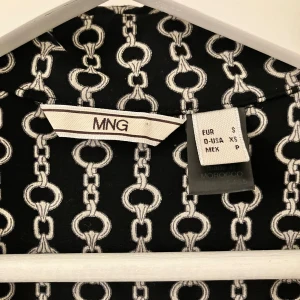 Svart långklänning från Mango  - Svart långklänning från MNG med vit kedjeinspirerat mönster. Klänningen har korta ärmar, v-ringad hals och knappar framtill. Materialet känns mjukt och följsamt, perfekt för dig som gillar coola grafiska prints och vill sticka ut.