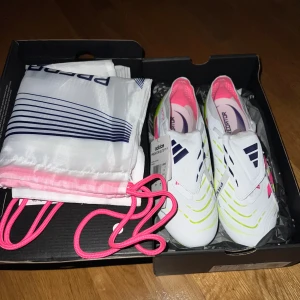 Adidas Predator Fotbollskor Elite  - Snygga Adidas Predator Elite FT fotbollsskor i vitt med neon-gula och rosa detaljer samt mörkblå logga. Skorna är aldrig använda och funkar på konst samt vanligt gräs. Skorna har en modern design med mönstrade ränder och texten Predator på sulan. Ovandelen är i syntetmaterial och sulan har fasta dobbar för gräset.