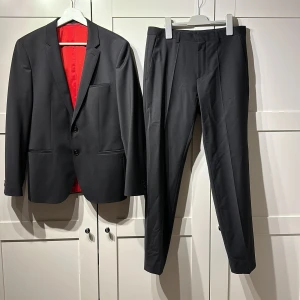 Svart kostym - HUGO ARTI HESTEN - Stilren svart kostym från Hugo Boss med klassisk kavaj och matchande kostymbyxor. Kavajen har två knappar, smal passform och röd insida med Hugo-logga. Byxorna är raka och har pressveck. Perfekt för dig som vill ha en snygg och modern look. Använd endast vid två tillfällen! 