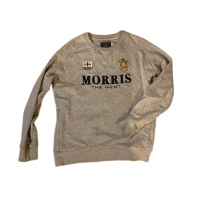 Grå sweatshirt från Morris Gents - Snygg grå sweatshirt från Morris Gents i 100% bomull. Tröjan har rund hals, broderade detaljer med brittisk flagga och emblem på bröstet samt stor Morris-logga. Perfekt för dig som gillar klassisk stil med en twist.