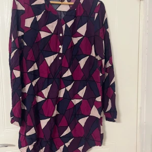 Färgstark blus från Jyothi med grafiskt mönster - Säljer en långärmad blus från Jyothi med ett coolt grafiskt mönster i lila, rosa och beige toner. Blusen har knappar framtill och är tillverkad i 100% viskos, vilket gör den mjuk och skön. Perfekt för dig som gillar att sticka ut med färg och form!