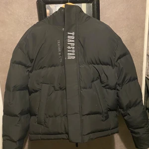 Svart pufferjacka från Trapstar - Trapstar pufferjacka i svart med broderad logga på ryggen och huvan, samt texten 'IT'S A SECRET'. Jackan har huva, två sidofickor och dragkedja framtill. Märkespatch på ärmen och coola detaljer som ger jackan en streetwear-vibe.