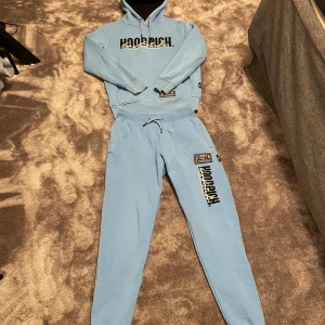 Hoodrich tracksuit - Säljer en ljusblå hoodie från Hoodrich med stor broderad logga på bröstet och tryck på ryggen. Hoodien har huva med snörning, ribbade muddar och en stor magficka. Materialet är mjuk bomull och polyester, perfekt för chill dagar.