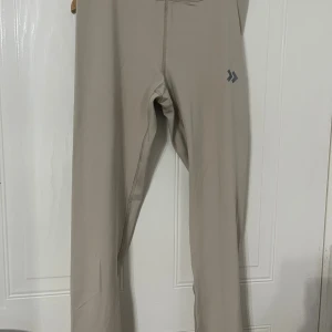 Beige träningsleggings från 157 - Säljer ett par beige träningsleggings från 157 med diskret logga på låret. Byxorna är tillverkade i ett stretchigt syntetmaterial med quickdry-teknologi, perfekt för träning. De har en tight passform och hög midja för extra komfort.