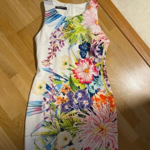 Vit blommig kortklänning från Adrianna Papell - Supersnygg vit kortklänning från Adrianna Papell med färgstarkt blommigt mönster framtill. Klänningen är ärmlös och har en figurnära passform med rund halsringning och dragkedja i ryggen. Perfekt för dig som gillar färg och vill sticka ut.