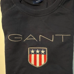Mörkblå GANT sweatshirt med emblem - Snygg mörkblå sweatshirt från GANT med broderad logga och ett stort emblem med stjärnor och rödvita ränder på bröstet. Rund hals med röd, vit och blå randig insida. Långärmad och tillverkad i mjuk bomull, perfekt för en chill och stilren look.