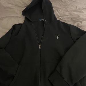 Svart hoodie från Polo Ralph Lauren - Svart hoodie från Polo Ralph Lauren med klassisk broderad logga på bröstet. Tröjan har huva, dragkedja framtill och är tillverkad i en mjuk blandning av bomull och återvunnen polyester. Perfekt för chill dagar och enkel att matcha med jeans eller joggers. Priset kan alltid sänka priset🤙🏾
