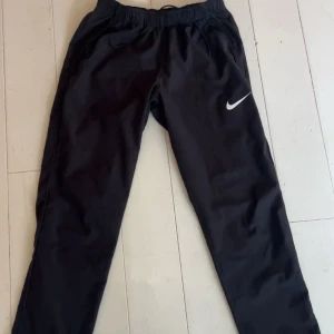 Svarta träningsbyxor från Nike - Svarta träningsbyxor från Nike med klassisk vit swoosh-logga på benet. Byxorna har resår och snörning i midjan för skön passform. Perfekta för träning eller chill, tillverkade i lätt och smidigt material. Knappt använda 