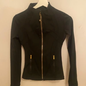 Lululemon define jacket - Säljer en svart, figursydd jacka med guldiga dragkedjor framtill och på fickorna från lululemon. Jackan har hög krage och långa ärmar, perfekt för en sportig och stilren look. Materialet känns mjukt och stretchigt, vilket gör den bekväm att bära. I stl 2/S men passar xs.