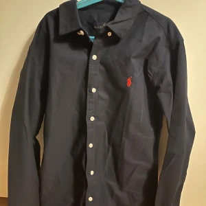 Mörkblå  skjorta från Ralph Lauren  - Klassisk mörkblå  skjorta från Ralph Lauren med röd broderad logga på bröstet. Skjortan har lång ärm, knappar framtill och är gjord i mjuk bomull. Perfekt för dig som gillar stilrena och tidlösa plagg med en sportig touch. Size S pris kan diskuteras fraktar samma dag ! 