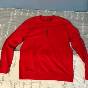 Röd sweatshirt från Hugo Boss - Röd sweatshirt från Hugo Boss med svarta detaljer längs ärmarna och ett tryckt logomönster på bröstet. Tröjan har rund halsringning och långa ärmar. Perfekt för dig som vill ha en stilren och trendig look.