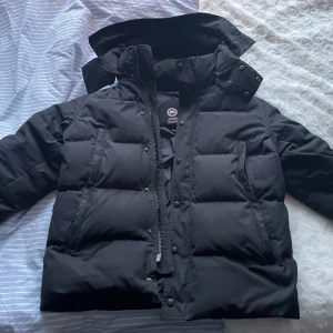 Svart wyndham från Canada Goose - Säljer en svart dunjacka från Canada Goose i storlek L. Jackan har en klassisk pufferstil med hög krage, dragkedja och tryckknappar framtill. Fylld med dun för extra värme och har en stilren look som passar perfekt till vintern. Jackan har några få defekter 