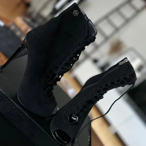 Svarta stövletter med klack från Philipp Plein - Säljer ett par svarta stövletter från Philipp Plein , med hög smal klack och snörning framtill. Skorna har öppen tå, dragkedja på sidan och snygga hjärtformade detaljer i metall. Tillverkade i mocka med glansiga inslag på klacken och tåpartiet. Dem har även fått en sula undertill så dem är klara att använda, använda 1 gång 