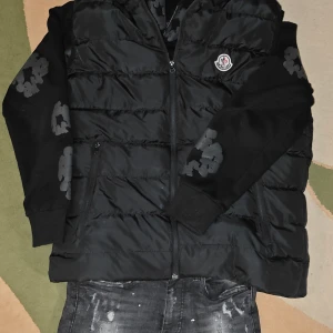 Svart dunväst från Moncler - Säljer en svart dunväst från Moncler med klassisk quiltad design och dragkedja framtill. Västen har Moncler-logga på bröstet och innerfoder med Moncler-tryck samt serietidningsetikett. Perfekt för lager-på-lager och streetstyle. Har fläckar och ett små hål