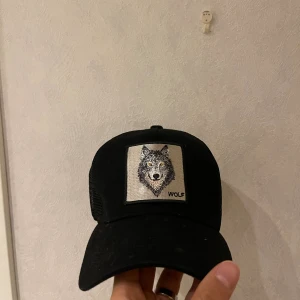 Svart truckerkeps med vargmärke - Svart truckerkeps med mesh på sidorna och justerbar snapback. Framtill finns ett broderat märke med en varg och texten 'WOLF'. Kepsen har böjd skärm och cool streetstil, perfekt för dig som gillar djurmotiv och vill sticka ut.