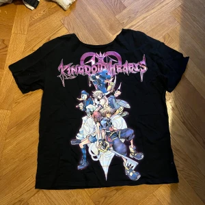 Kingdom Hearts svart t-shirt - Svart t-shirt med stort färgglatt Kingdom Hearts-tryck på ryggen. Motivet visar flera karaktärer från spelet och loggan i lila och rosa. T-shirten har klassisk passform och är kortärmad, perfekt för dig som älskar gaming och anime.