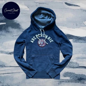Blå hoodie från Abercrombie & Fitch - Snygg marinblå hoodie från Abercrombie & Fitch med broderad logga och text på bröstet. Tröjan har huva med snörning, känguruficka och ribbade muddar. Insidan av huvan är fodrad i grått tyg för extra komfort. Perfekt för chill dagar.