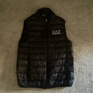  EA7 Emporio Armani väst svart - Snygg svart dunväst från EA7 Emporio Armani med quiltad design och logotyp på bröstet. Västen har dragkedja med EA7-detalj och står ut med sin stilrena look. Perfekt för dig som vill ha en modern och sportig vibe.