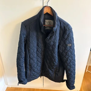 Mörkblå quiltad jacka från Cutter & Buck - Snygg mörkblå quiltad jacka från Cutter & Buck med dragkedja och hög krage. Jackan har två fickor med dragkedja och ett diskret logomärke på ärmen. Perfekt för kyliga dagar och har en stilren look med rutmönstrad struktur.