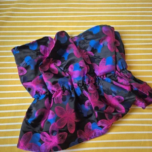 Blommig bandeau tubtopp i satin - Snygg bandeau tubtopp i glansig satin med blommigt mönster i lila, rosa och blått på svart botten. Toppen har en figurnära passform och är perfekt för dig som vill sticka ut med färg och mönster.