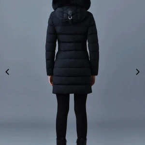 Svart lång dunjacka med pälskant - Snygg svart dunjacka från Mackage med lång passform, stor huva med fluffig pälskant och tvåvägsdragkedja framtill. Jackan har quiltad design, långa ärmar och praktiska fickor med dragkedja. Perfekt för kalla vinterdagar. Storlek XS. Inköpspris: 14000kr. Säljer nu pga för liten. 